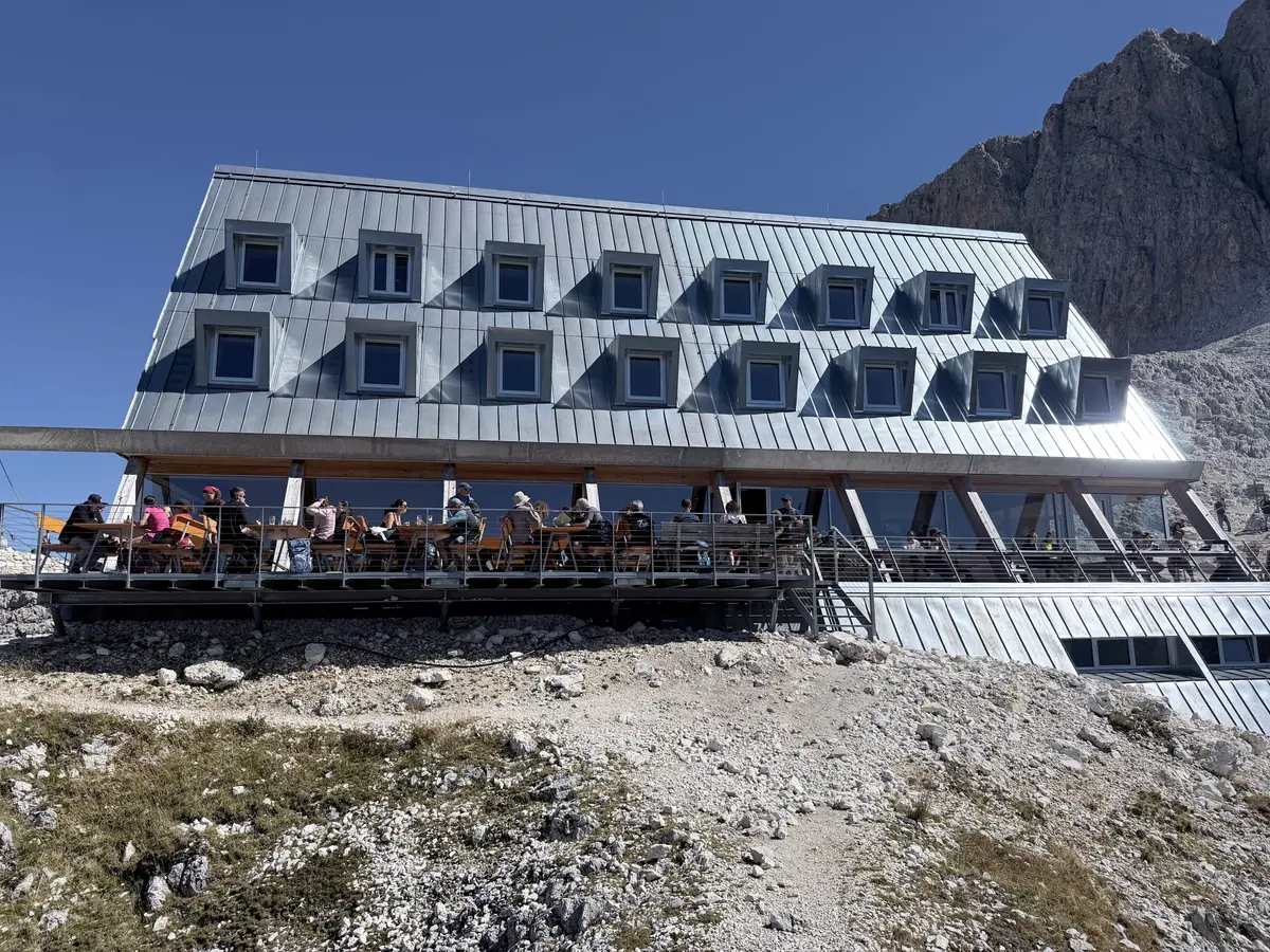 金屬外牆的現代建築玫瑰山屋，一樓露台坐滿健行者，背景是晴朗藍天與岩峰