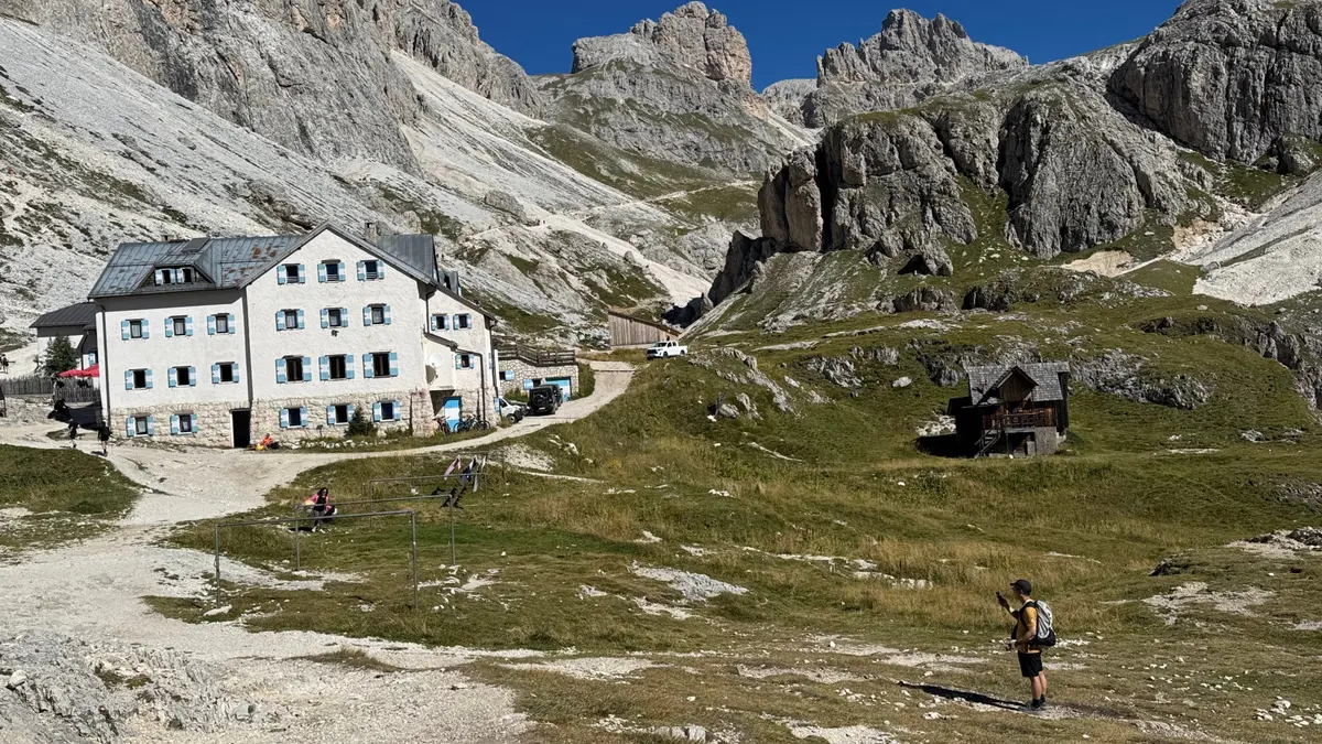 白色多層的 Rifugio Vajolet 與旁邊較小的 Rifugio Preuss，背後是鋸齒狀岩峰，健行者走在草坡上
