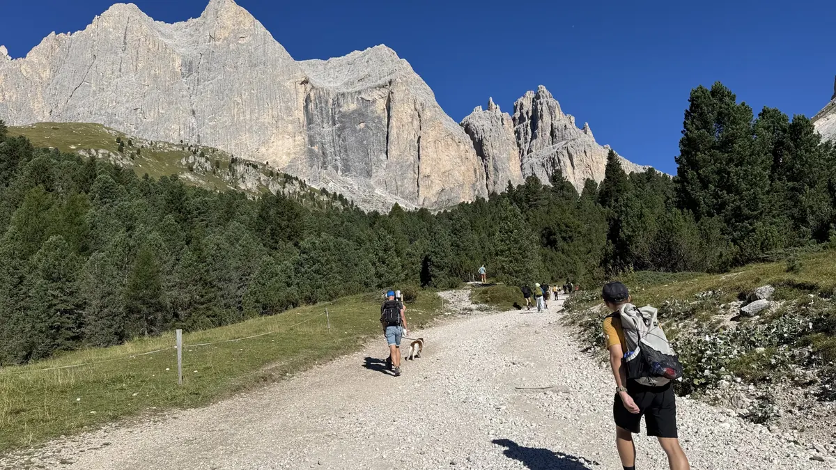 Gardeccia 之後的寬闊碎石步道，健行者往岩壁方向前進，兩側是草地與松樹