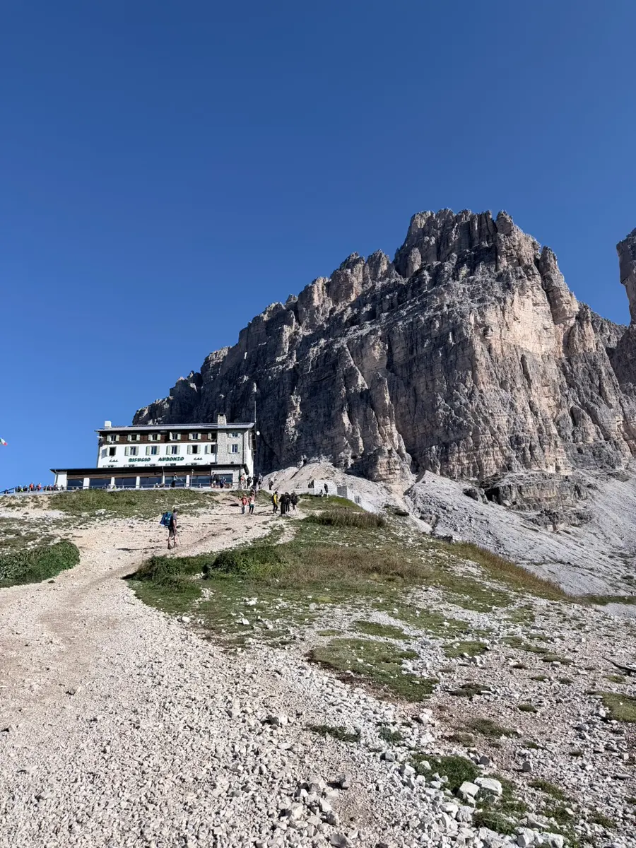 藍天下的 Rifugio Auronzo 山屋，建築後方是高聳的灰白色岩壁，前方碎石坡上有登山客往上走