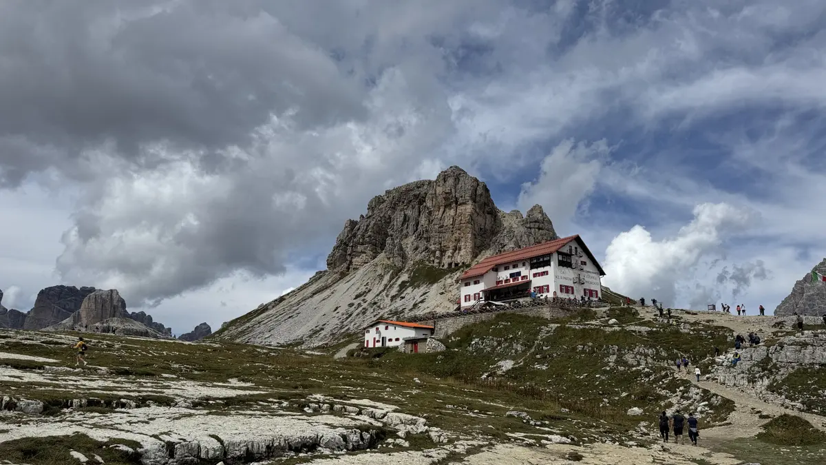Locatelli 山屋（Drei Zinnen Hütte）白牆紅屋頂的建築，後方是岩峰，步道上有登山客往山屋走