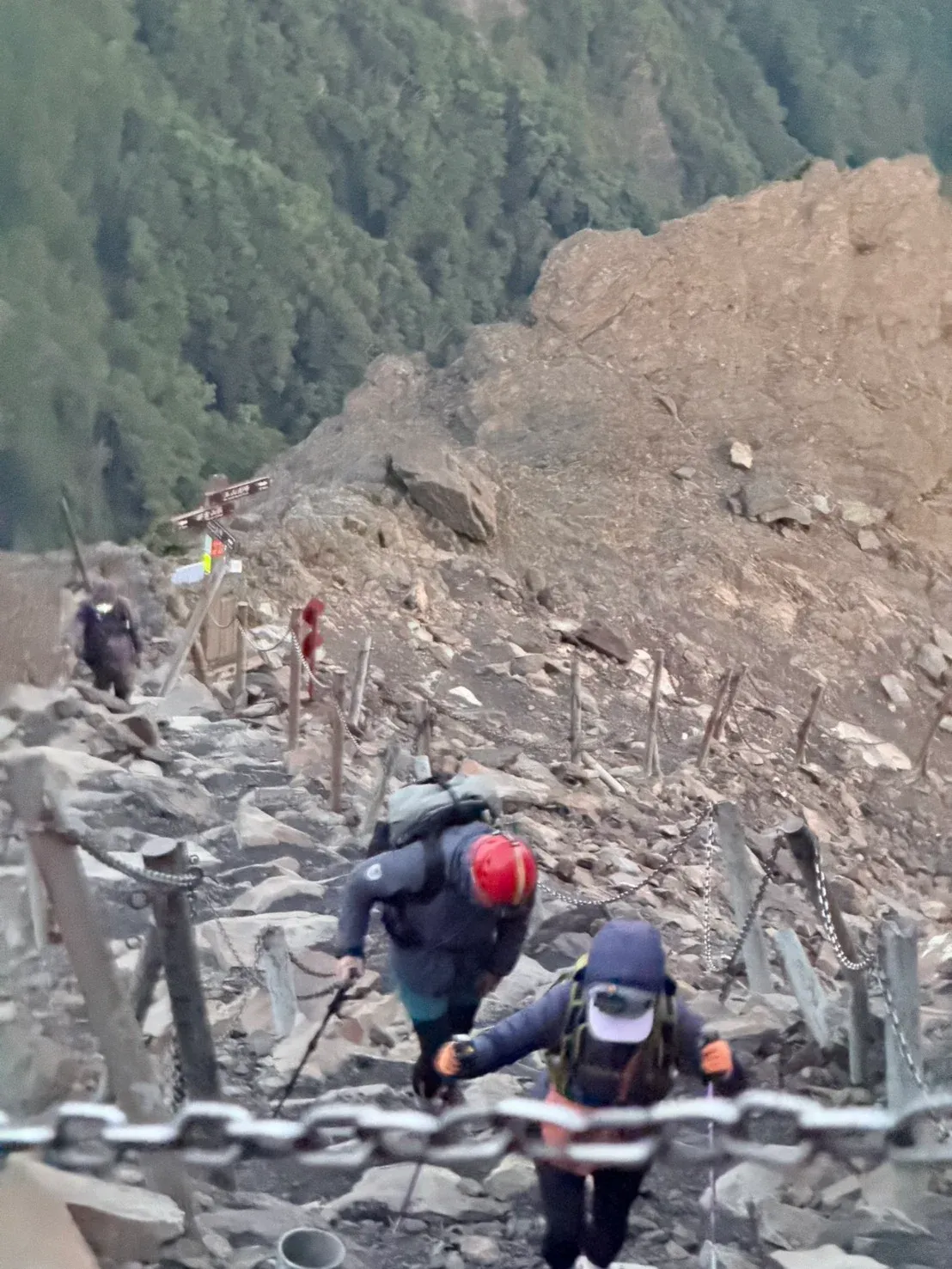 玉山主峰碎石坡上的清晨，山友穿著外套、戴帽子和岩盔，沿鐵鍊護欄往上走