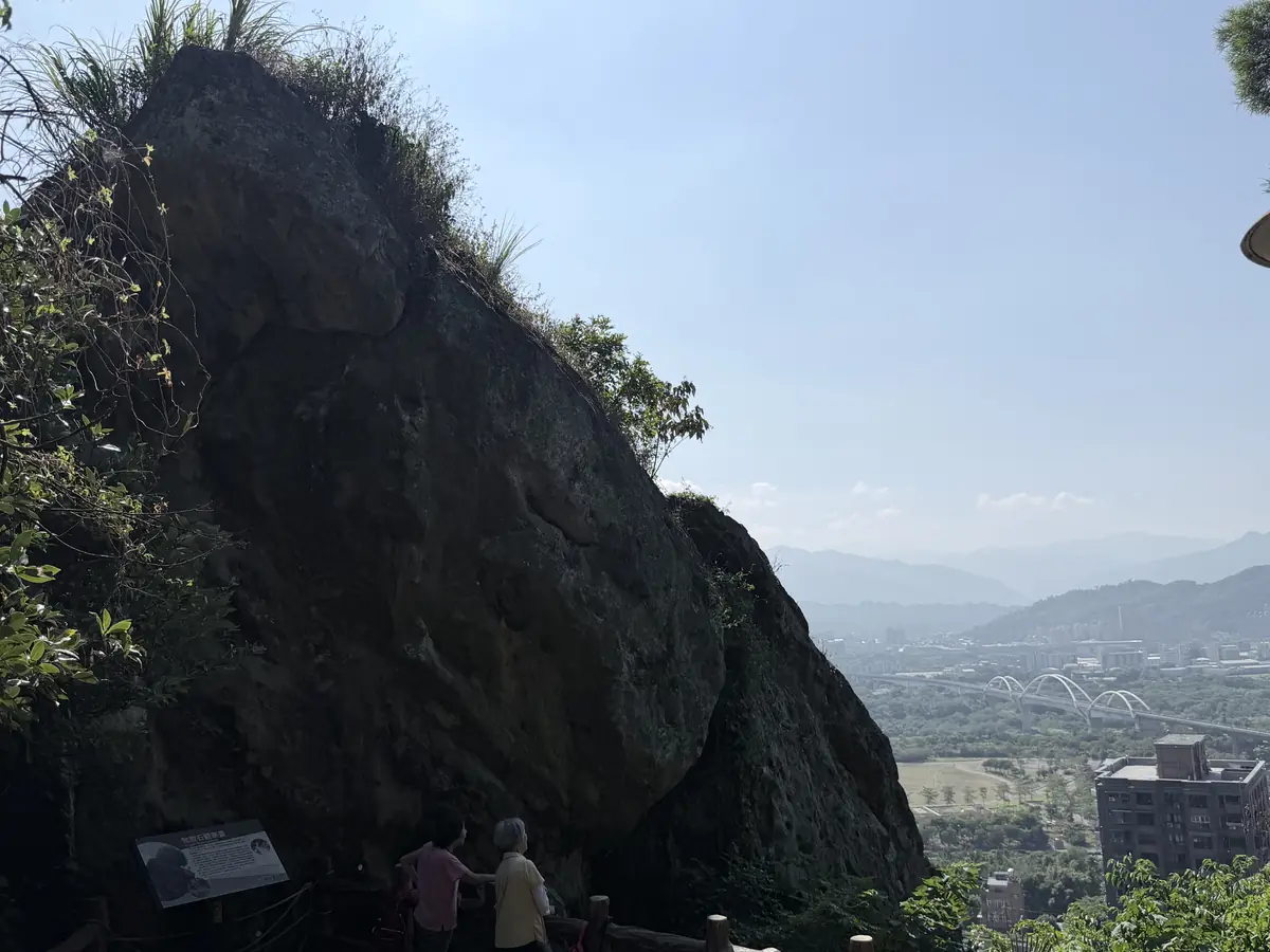 近看鶯歌石的深色岩壁，巨岩幾乎佔滿畫面，右側可以看到遠方的山稜和市區